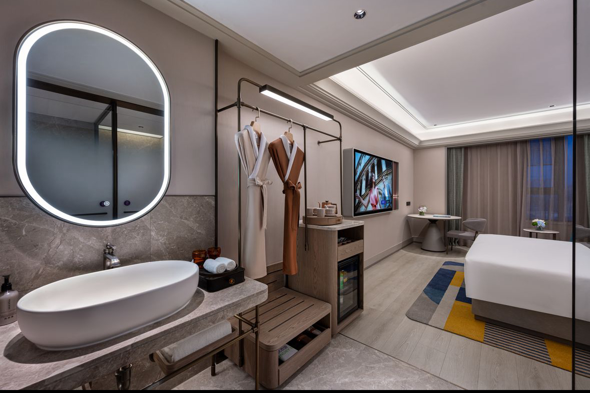 Modern hotel room with illuminated mirror, vanity, bed, and seating area.