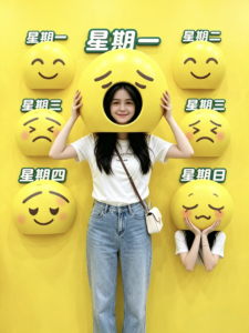 A visitor posing in a yellow weekday-themed photo wall with expressive emoji faces.