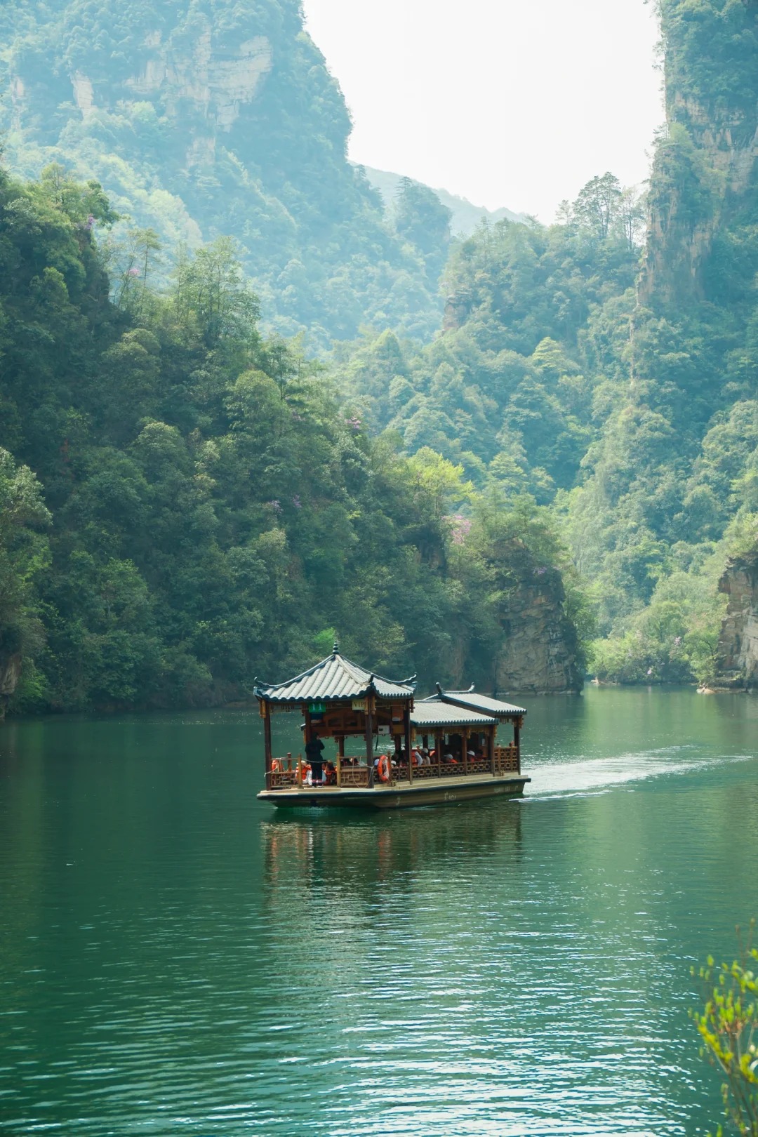 A covered sightseeing boat crossing Baofeng Lake beneath forested sandstone cliffs.