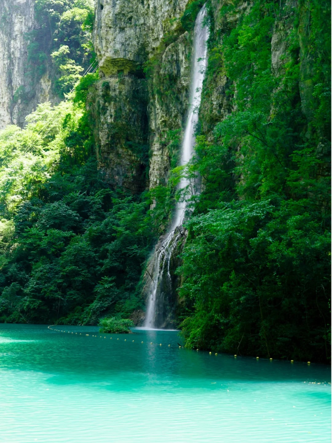 Tall narrow waterfall dropping from a forested cliff into turquoise water at Baofeng Lake.