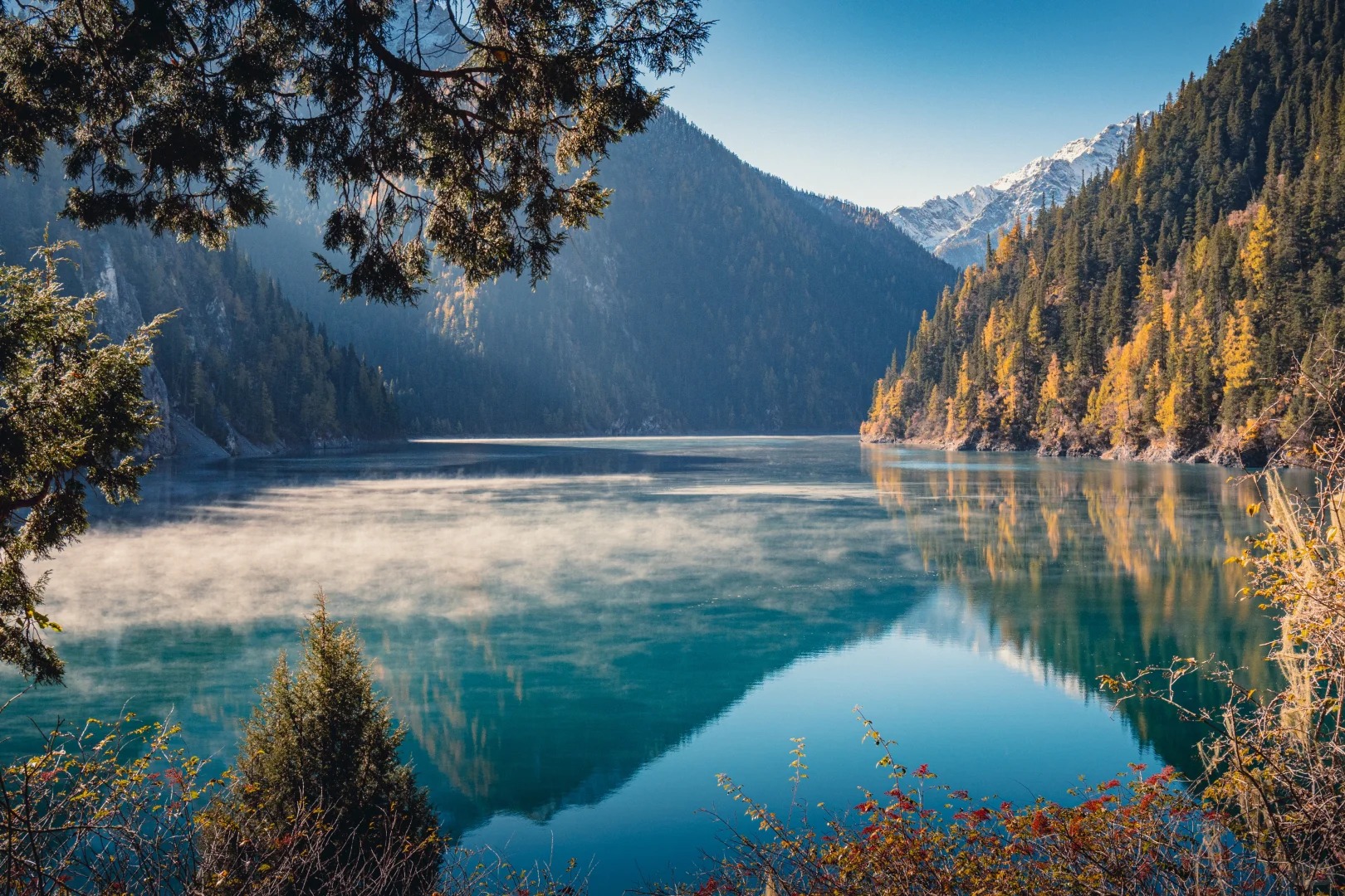 Long Lake in Jiuzhaigou with forested mountains, distant snow peaks and light mist.